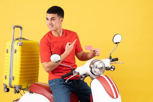 Vorderansicht lächelnder junger Mann im roten T-Shirt auf Moped, das Kreditkarte hält
