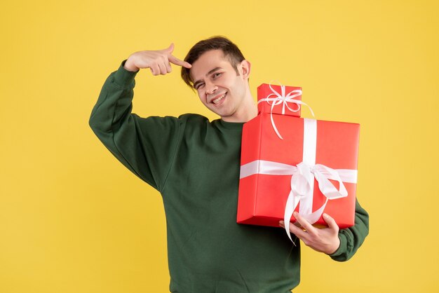 Vorderansicht lächelte junger Mann mit Weihnachtsgeschenk, das auf Geschenke zeigt, die auf Gelb stehen