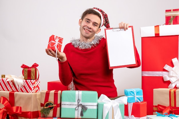 Vorderansicht lächelte junger Mann mit Weihnachtsmütze, der um Weihnachtsgeschenke sitzt