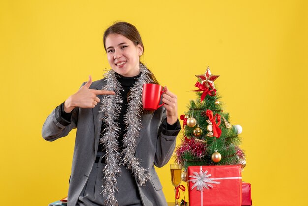 Vorderansicht lächelte junges Mädchen, das rote Tasse hält, die mit Finger selbst nahe Weihnachtsbaum und Geschenkcocktail zeigt