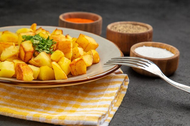 Vorderansicht leckere Bratkartoffeln im Teller mit Gewürzen auf einem dunkelgrauen Tisch