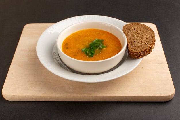 Vorderansicht leckere Gemüsesuppe in Teller mit Brotlaib auf dunklem Schreibtisch.