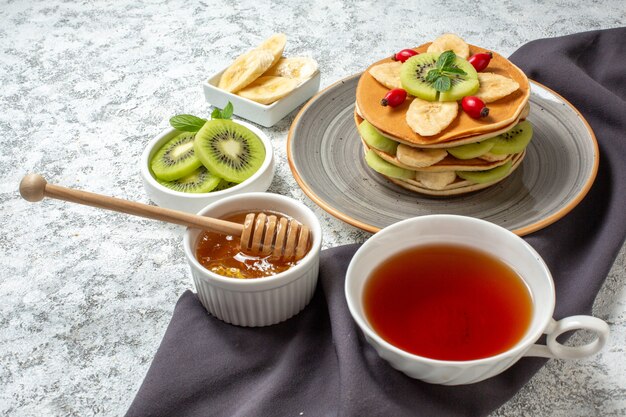 Vorderansicht leckere Pfannkuchen mit geschnittenen Früchten und Tasse Tee auf weißer Oberfläche Obst süße Dessert Frühstück Farbe Kuchen Zucker