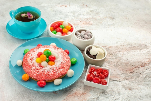 Vorderansicht leckerer rosa Kuchen mit bunten Bonbons und Tasse Tee auf weißem Hintergrund Dessertkuchen Regenbogenfarben Kuchen Süßigkeiten