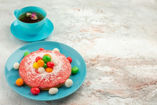 Vorderansicht leckerer rosa Kuchen mit bunten Bonbons und Tasse Tee auf weißem Hintergrund Kuchen Regenbogenfarben Kuchen Dessert Süßigkeiten can