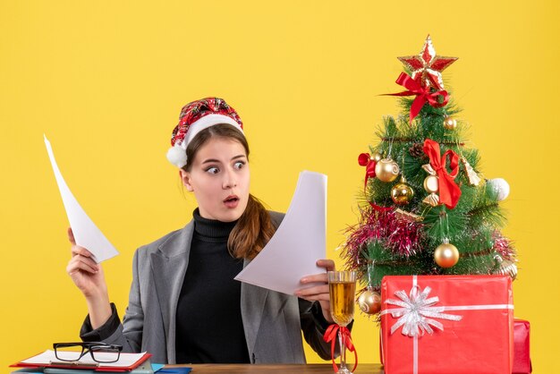 Vorderansicht-Mädchen mit großen Augen und Weihnachtsmütze, die am Tisch sitzen und Dokumente Weihnachtsbaum und Geschenkcocktail betrachten