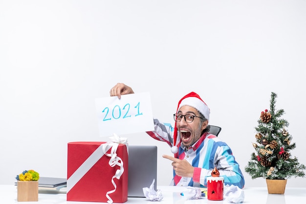 Vorderansicht männlicher Arbeiter, der an seinem Arbeitsplatz sitzt und Papierblatt mit der Nummer 2021 hält, Neujahrskonzept Business Job Weihnachtsbüro