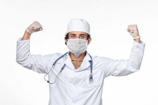 Vorderansicht männlicher Arzt im medizinischen Anzug, der sterile Maske als Schutz vor Covid Flexing auf White Wall Splash Virus Coronavirus Pandemie Gesundheit trägt