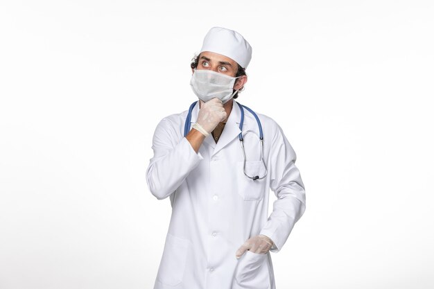 Vorderansicht männlicher Arzt im medizinischen Anzug mit Maske als Schutz vor kovidem Denken auf Pandemiemedizin des Coronavirus-Virus mit weißer Wand
