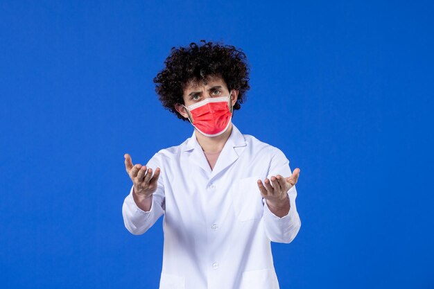 Vorderansicht männlicher Arzt im medizinischen Anzug mit roter Maske auf blauem Hintergrund Drogenvirus Covid-Impfstoff Gesundheitsmedizin Krankenhauspandemie