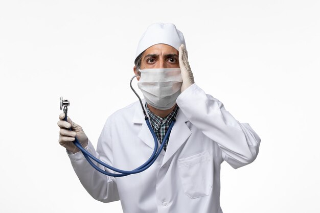 Vorderansicht männlicher Arzt im medizinischen Anzug und in der Maske wegen Coronavirus, der Stethoskop auf Weißbodenvirus-Pandemie-Isolationskrankheit hält covid-