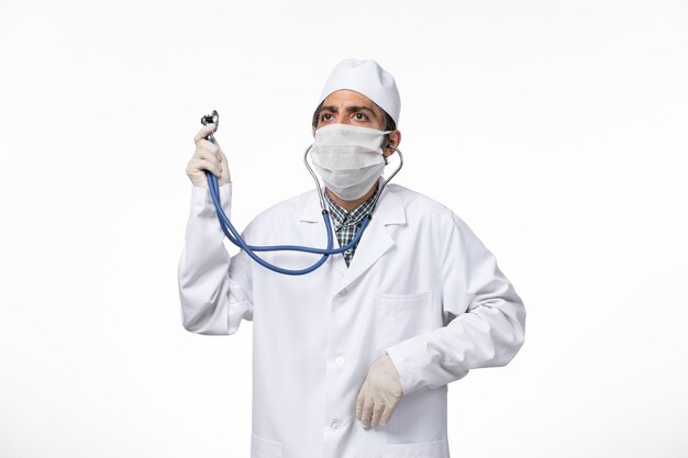 Vorderansicht männlicher Arzt im medizinischen Anzug und in der Maske wegen Coronavirus unter Verwendung seines Stethoskops auf weißer Oberfläche