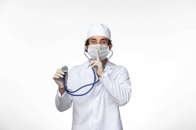 Vorderansicht männlicher Arzt im medizinischen Anzug und Tragen einer Maske als Schutz vor Covid - unter Verwendung eines Stethoskops bei hellweißer Wandviruskrankheit Covidpandemie