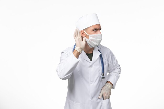 Vorderansicht männlicher Arzt im medizinischen Anzug und Tragen einer Maske gegen Covid mit blauem Stethoskop, das versucht, auf weißer Wand Covid Disease Disease Pandemic Virus zu hören