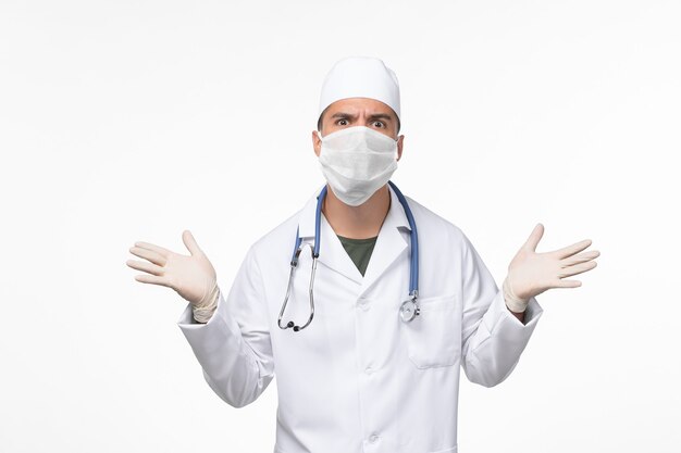 Vorderansicht männlicher Arzt im medizinischen Anzug und Tragen einer Maske gegen Covid mit Stethoskop auf hellweißer Wand Covid-Disease-Pandemie