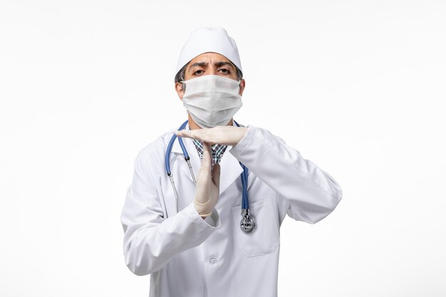 Vorderansicht männlicher Arzt im weißen medizinischen Anzug mit Maske wegen Covid auf hellweißer Oberfläche