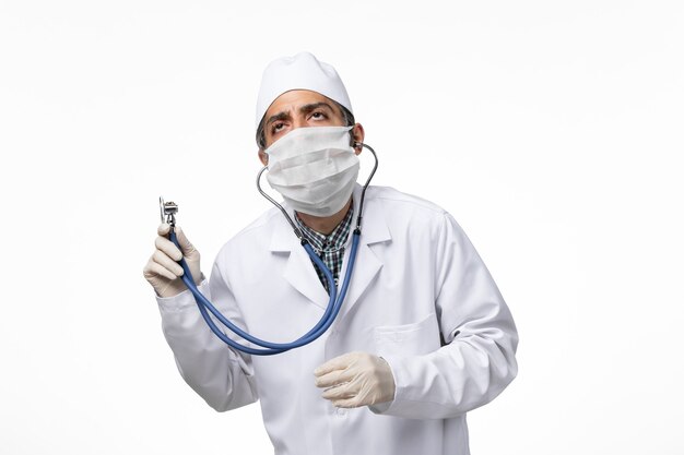 Vorderansicht männlicher Arzt im weißen medizinischen Anzug und in der Maske aufgrund des Coronavirus auf der weißen Oberfläche