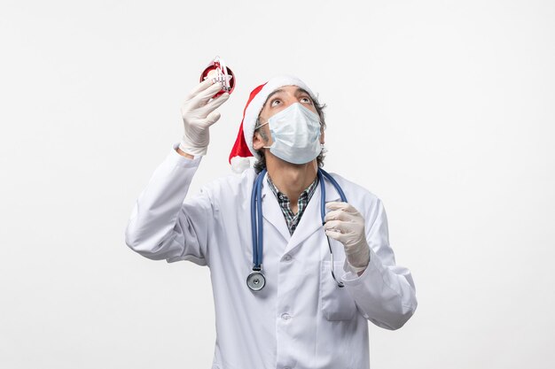 Vorderansicht männlicher Arzt in Maske mit Spielzeug auf weißem Wandgesundheits-Covid-Feiertagsvirus
