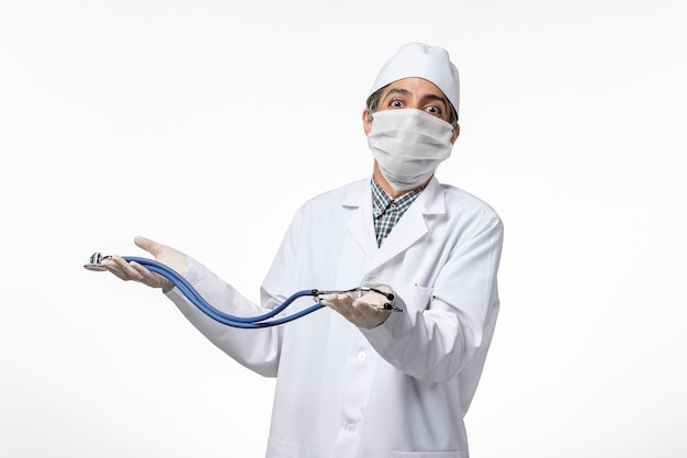 Vorderansicht männlicher Arzt in medizinischem Anzug und Maske aufgrund von Coronavirus unter Verwendung eines Stethoskops auf hellweißer Oberfläche