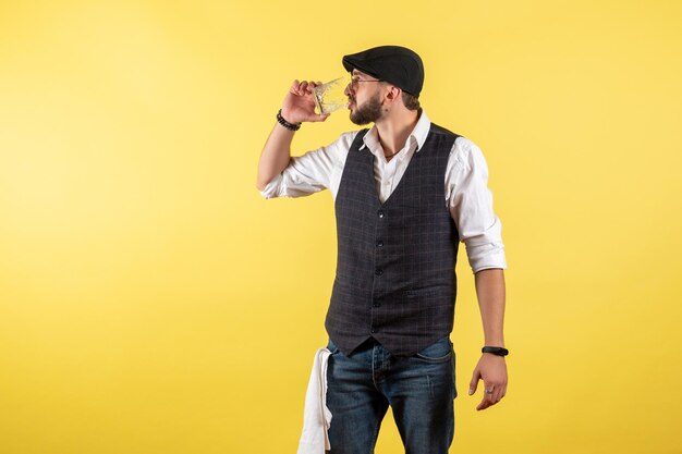 Vorderansicht männlicher Barkeeper, der aus Glas auf gelber Wand trinkt, trinken Alkohol Job Club Bar Nacht männlich