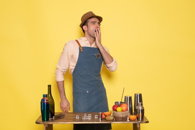 Vorderansicht männlicher Barkeeper, der vor dem Schreibtisch mit Getränken und Früchten steht, die auf gelber Wand gähnen, Getränkebar-Nachtclubsaft