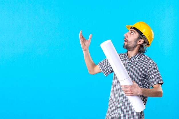 Vorderansicht männlicher Baumeister in gelbem Helm mit Plan auf blauem Hintergrund Arbeiter Architektur Konstrukteur Gebäude Farbe Wohnung