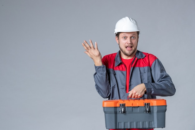 Vorderansicht männlicher Baumeister in Uniform mit Werkzeugkoffer auf grauem Hintergrund