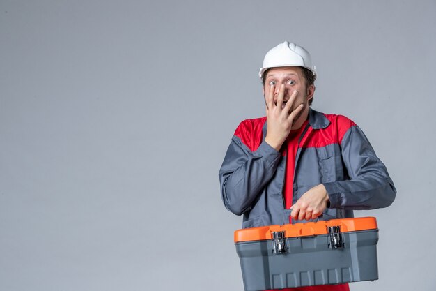 Vorderansicht männlicher Baumeister in Uniform mit Werkzeugkoffer überrascht und aufgeregt auf grauem Hintergrund