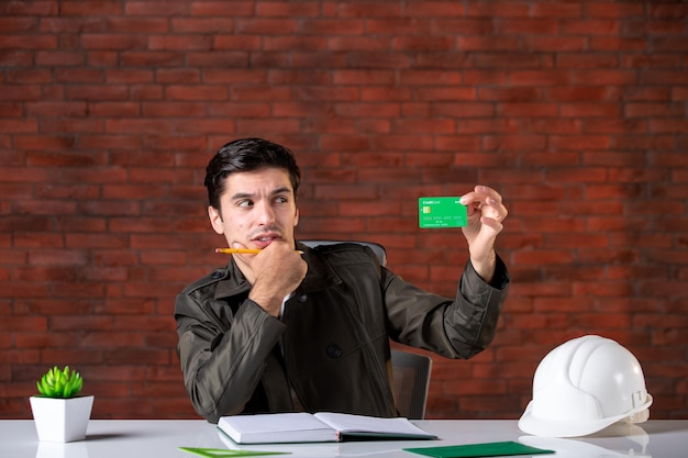 Vorderansicht männlicher ingenieur, der hinter seinem arbeitsplatz sitzt und grüne kreditkarten-ersteller-geldplan-unternehmens-geschäftsagenda hält