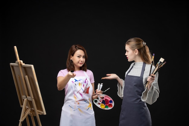 Vorderansicht malerinnen, die farben und quasten zum zeichnen auf schwarzer wand halten, zeichnen malerei-job-bilder kunstfoto-farbkünstler