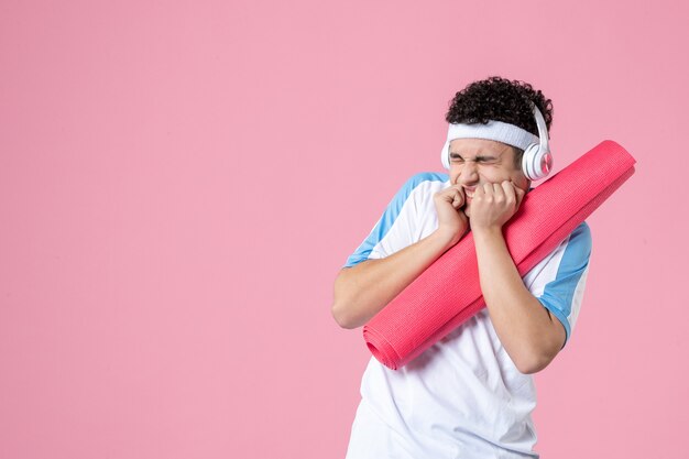Vorderansicht nervöser junger Mann in Sportkleidung mit Yogamatte und Kopfhörern