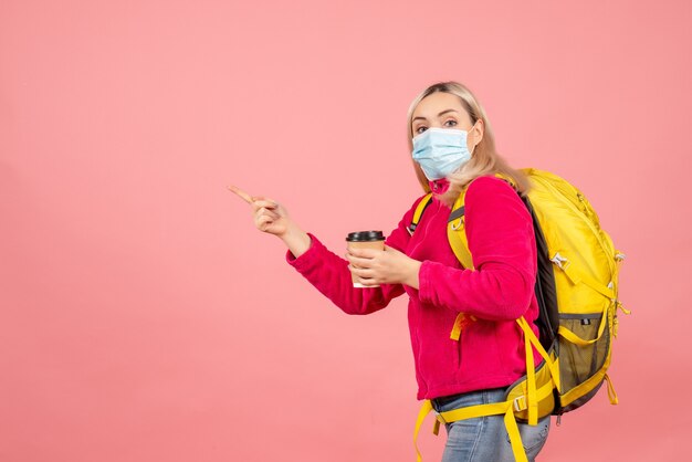 Vorderansicht reisende Frau mit gelbem Rucksack, der Maske hält, die Kaffeetasse hält, die nach links zeigt