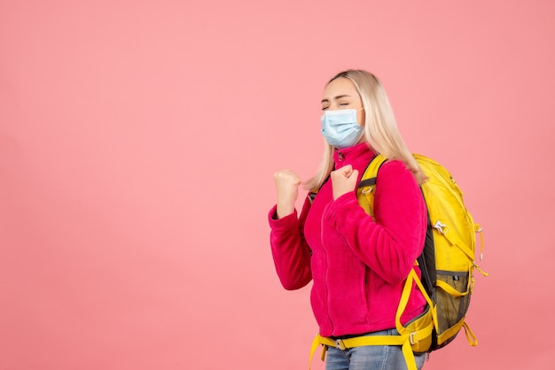 Vorderansicht reisende Frau mit gelbem Rucksack, der Maske trägt, die gewinnende Geste mit geschlossenen Augen zeigt