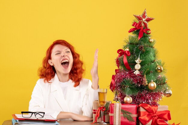 Vorderansicht-Ärztin, die hinter Tisch mit Weihnachtsgeschenken sitzt und ihre Hand auf gelbem Hintergrund erhebt