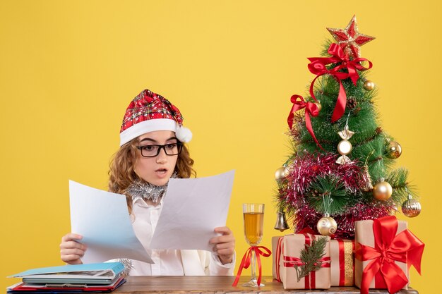 Vorderansicht-Ärztin, die mit Weihnachten sitzt, präsentiert Baum und hält Dokumente auf gelbem Hintergrund