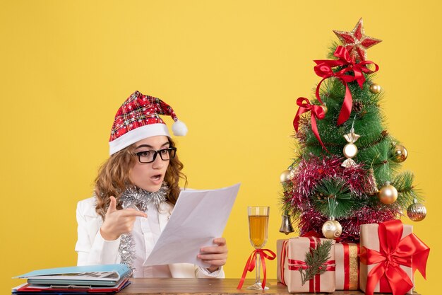 Vorderansicht-Ärztin, die mit Weihnachten sitzt, präsentiert Baum und hält Dokumente auf gelbem Hintergrund