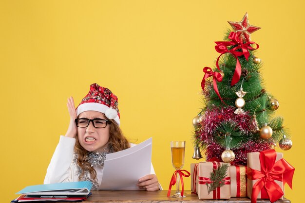 Vorderansicht-Ärztin, die mit Weihnachten sitzt, präsentiert Baum und hält Dokumente auf gelbem Hintergrund