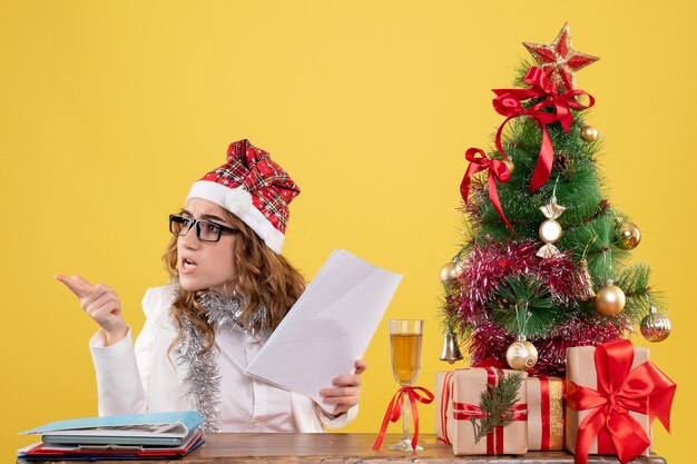 Vorderansicht-Ärztin, die mit Weihnachten sitzt, präsentiert Baum und hält Dokumente auf gelbem Hintergrund