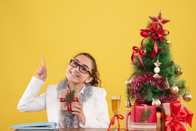 Vorderansicht-Ärztin, die mit Weihnachtsgeschenken und Baum auf gelbem Hintergrund sitzt