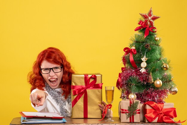 Vorderansicht-Ärztin, die mit Weihnachtsgeschenken und Baum auf gelbem Hintergrund sitzt