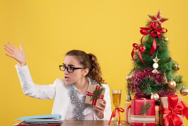 Vorderansicht-Ärztin, die mit Weihnachtsgeschenken und Baum auf gelbem Hintergrund sitzt