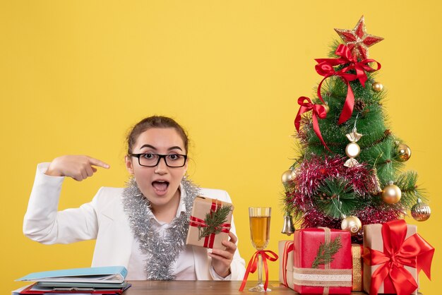 Vorderansicht-Ärztin, die mit Weihnachtsgeschenken und Baum auf gelbem Hintergrund sitzt