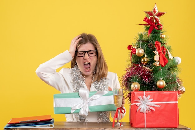 Vorderansicht-Ärztin, die vor Tisch mit Geschenken und Weihnachtsbaum auf gelbem Hintergrund sitzt