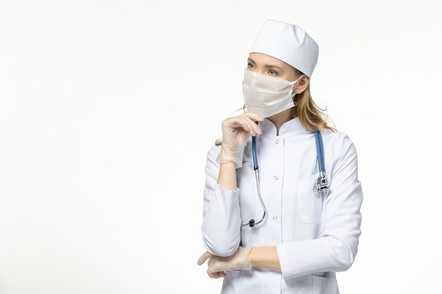 Vorderansicht Ärztin im medizinischen Anzug mit Maske und Handschuhen aufgrund des Coronavirus-Denkens über das Pandemie-Covid-Virus der hellweißen Wandkrankheit