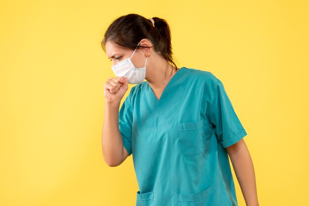 Vorderansicht Ärztin im medizinischen Hemd mit steriler Maske, die auf gelbem Hintergrund hustet