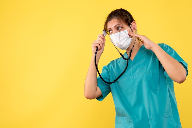 Vorderansicht-Ärztin im medizinischen Hemd und in der Maske mit Stethoskop auf gelbem Hintergrund