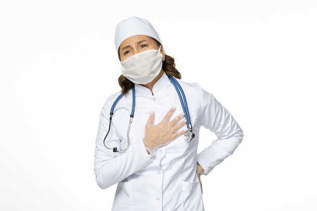 Vorderansicht Ärztin im weißen medizinischen Anzug und in der Maske, die Herzschmerz auf Weißwandpandemyvirus-Krankheitskrankheitsmedizin hat