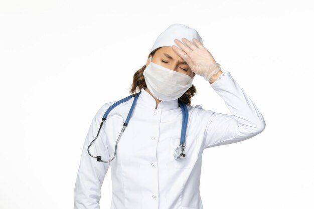 Vorderansicht Ärztin im weißen medizinischen Anzug und in der Maske, die Kopfschmerzen auf der weißen Wand-Pandemievirus-Krankheitskrankheitsmedizin hat