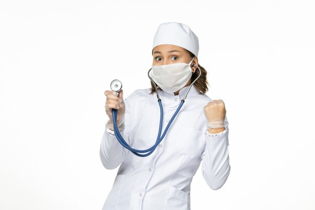 Vorderansicht Ärztin im weißen medizinischen Anzug und in der Maske unter Verwendung des Stethoskops auf der weißen Wandpandemieviren-Krankheitsmedizin