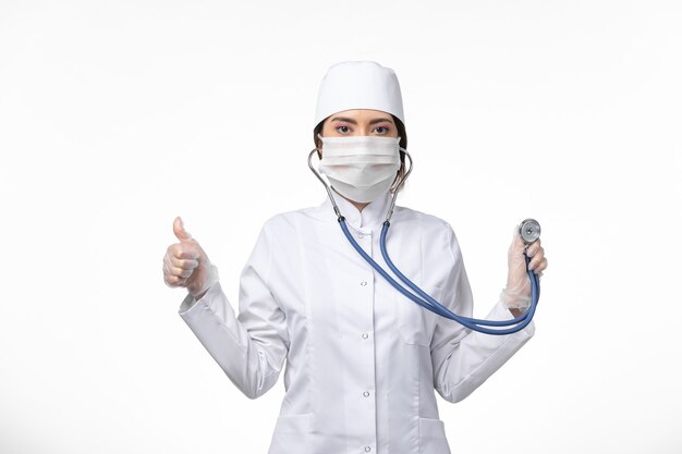 Vorderansicht Ärztin im weißen sterilen medizinischen Anzug und mit Maske aufgrund von Covid-Stethoskop auf White-Desk-Disease-Virus-Covid-Pandemie-Krankheit